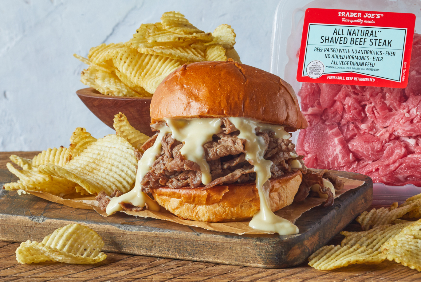 Trader Joe's All Natural Shaved Beef Steak; shown prepared in a sandwich with Cheddar Melange cheese sauce dripping from the top; Ghost Pepper Potato Chips surrounding and in background