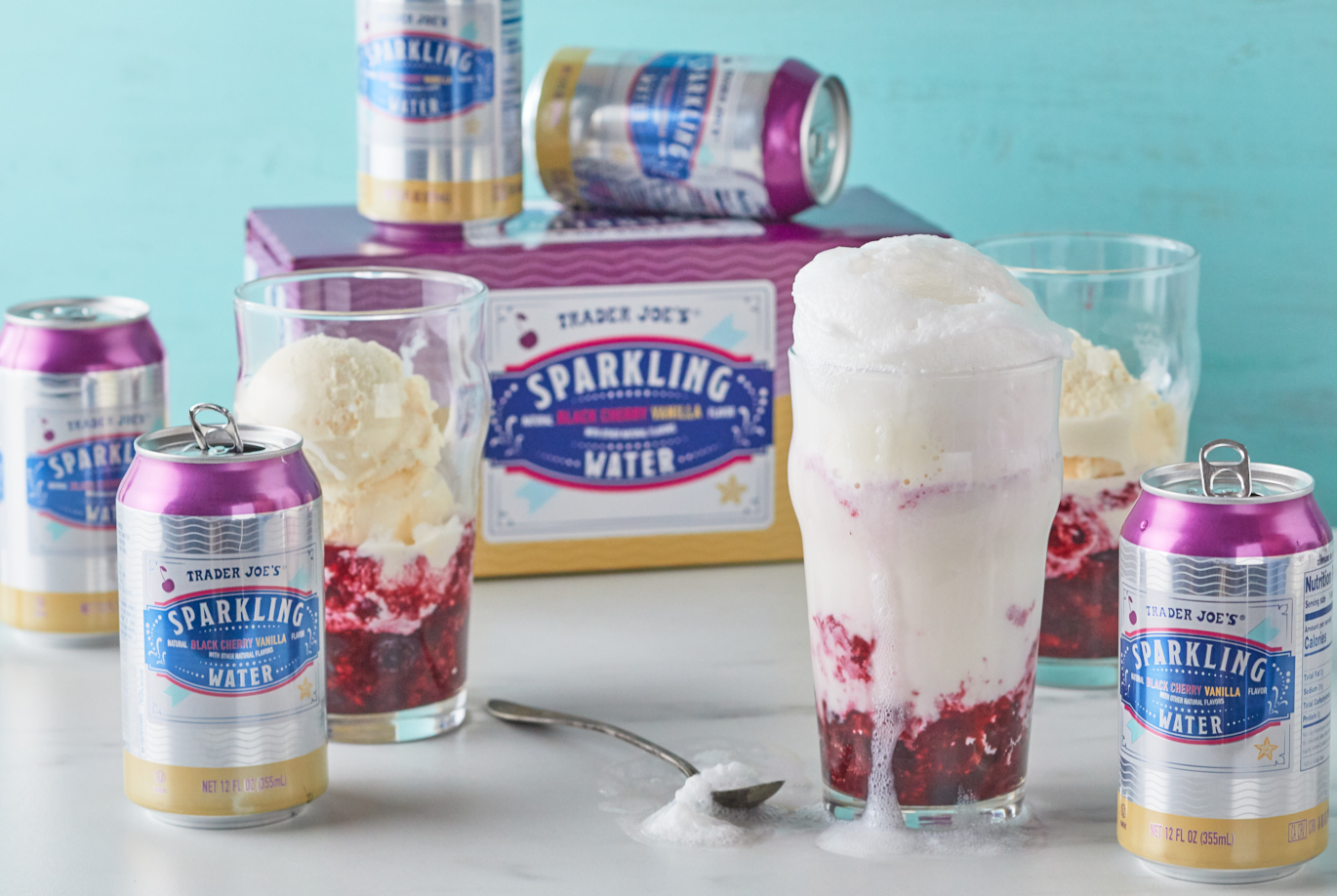 Trader Joe's Black Cherry Vanilla Flavored Sparkling Water; shown in a glass with ice cream and Verry Cherry Berry Blend, to make a recipe of Cherry Vanilla Ice Cream Float; blue background with more cans and glasses