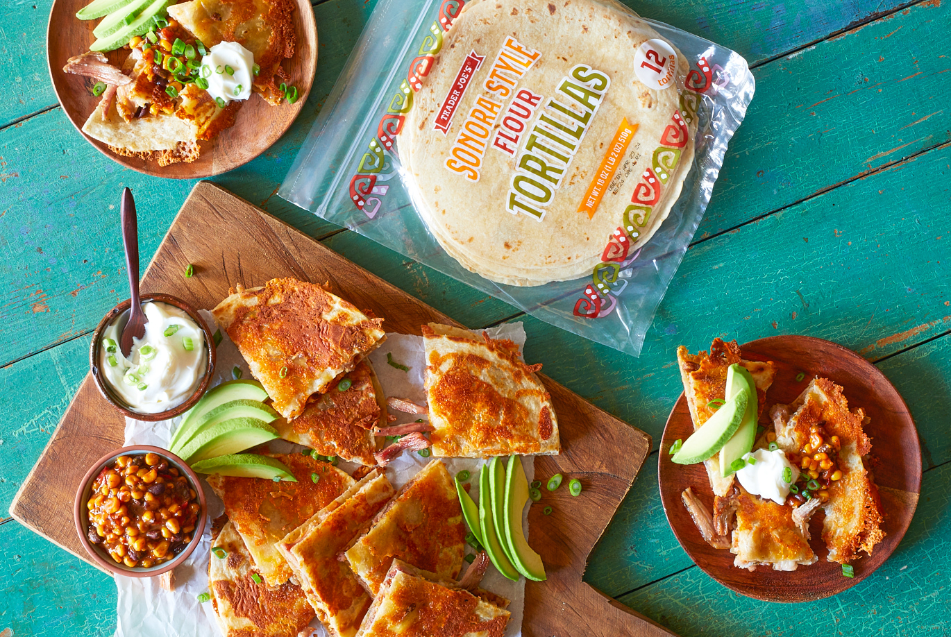 Trader Joe's Sonora Style Flour Tortillas; shown in recipe for crispy carnitas quesadillas; on green wood surface, several quesadillas cut in triangles and served with sliced avocado, Cowboy Quinoa Salsa, and sour cream