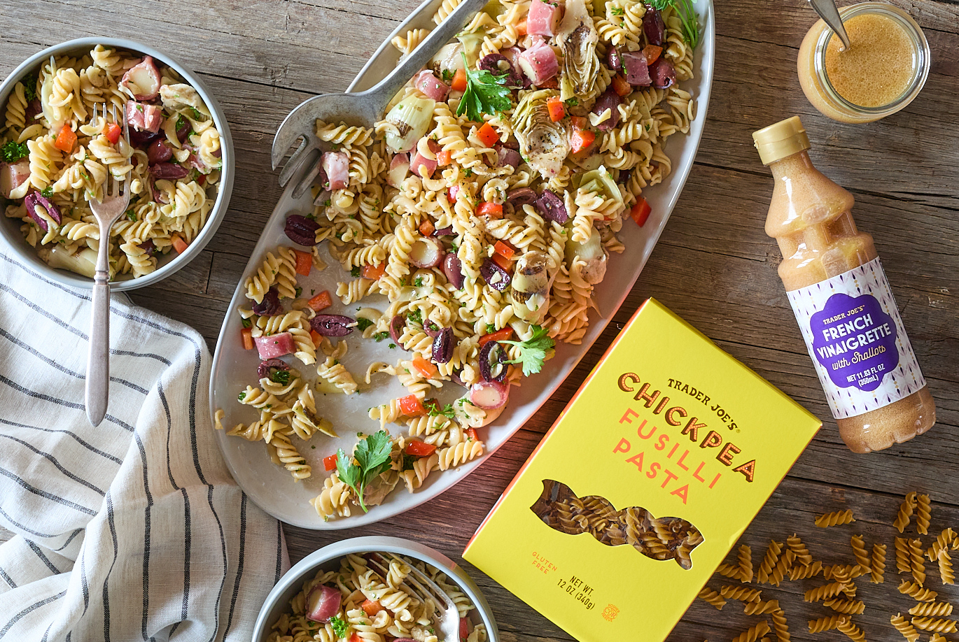 Trader Joe's Chickpea Fusilli Pasta and French Vinaigrette, shown in recipe for 'Antipasto Pasta Salad'. Serving platter and two bowls of Antipasto Pasta Salad, on top of a rustic wood surface. Dried pasta and jar of vinaigrette on right side