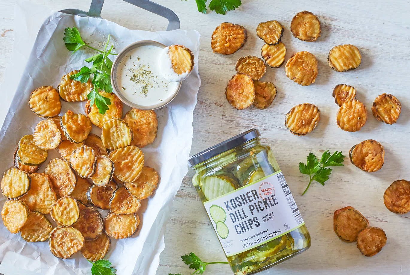 Trader Joe's Kosher Dill Pickle Chips used in recipe for 'Fried Pickle Chips'; on a silver tray with parchment papger, a side dish of ranch dressing and parsley garnish