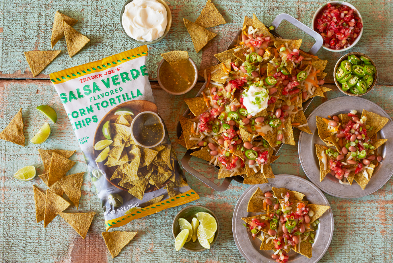 Trader Joe's Salsa Verde Flavored Corn Tortilla Chips prepared in a dish as nachos, with pinto beans, cheee, salsa, jalapeño slices and sour cream; plates and chips surrounding