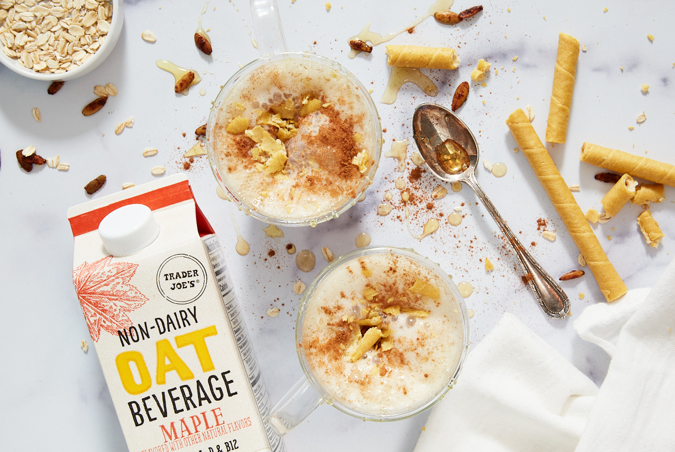 Trader Joe's Maple Non-Dairy Oat Beverage used in Maple Oat Latte recipe; in two glass mugs, with oats, cinnamon and maple surup scattered on surface