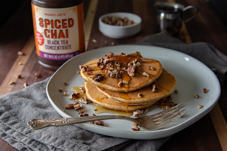 stack of spiced chai pancakes on plate topped with pecan pieces and syrup, next to a bottle of Trader Joe's Spiced Chai Black Tea Concentrate