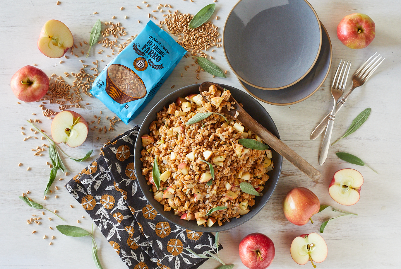 Trader Joe's 10 Minute Farro shown in recipe for Farro with Chicken Sausage and Apples; white surface and black serving bowl with Farro; dry grains sprinkled around, with apples and sage garnish