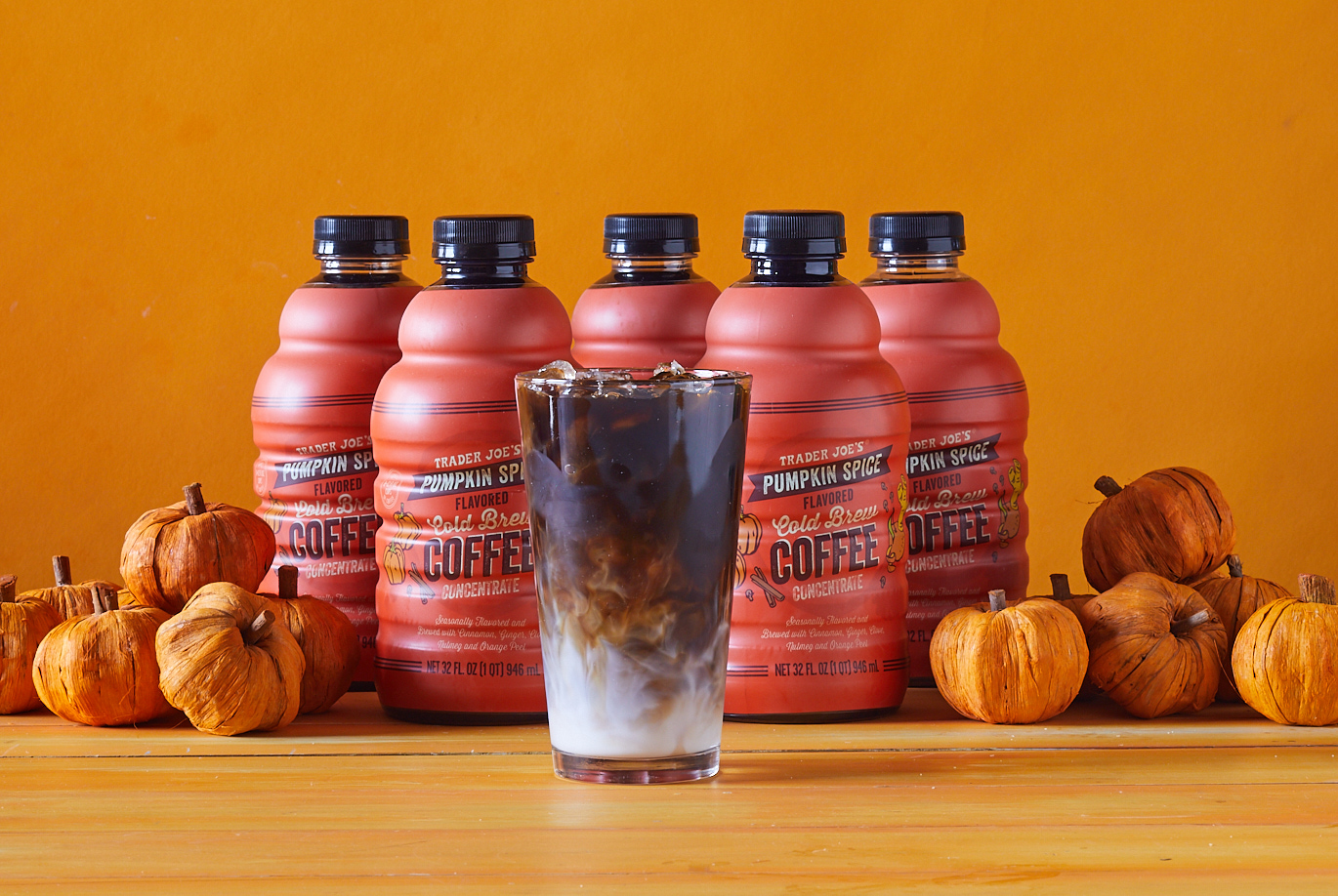 Trader Joe's Pumpkin Spice Cold Brew Coffee; orange surface and orange background; several bottles of Cold Brew lined up, with a glass full of iced cold brew and a swirl of milk at the bottom; decorative pumpkins surrounding