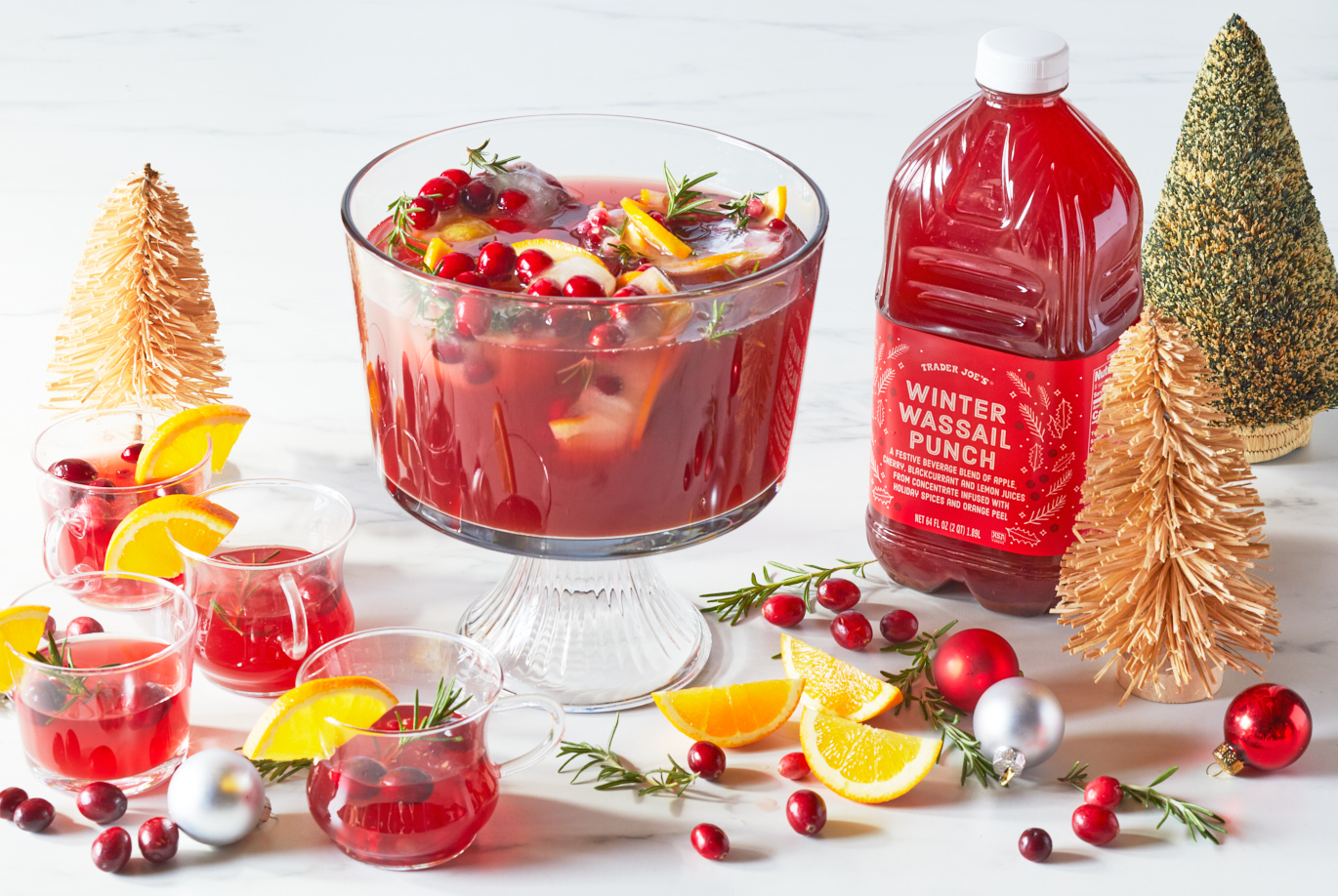 Trader Joe's Winter Wassail Punch; in a punch bowl, surrounded by festive glasses filled with punch, cranberries and rosemary garnishes