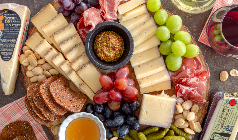 cheese board covered with piles of grapes, crackers, mustard, honey, prosciutto, cornichons, and almonds.