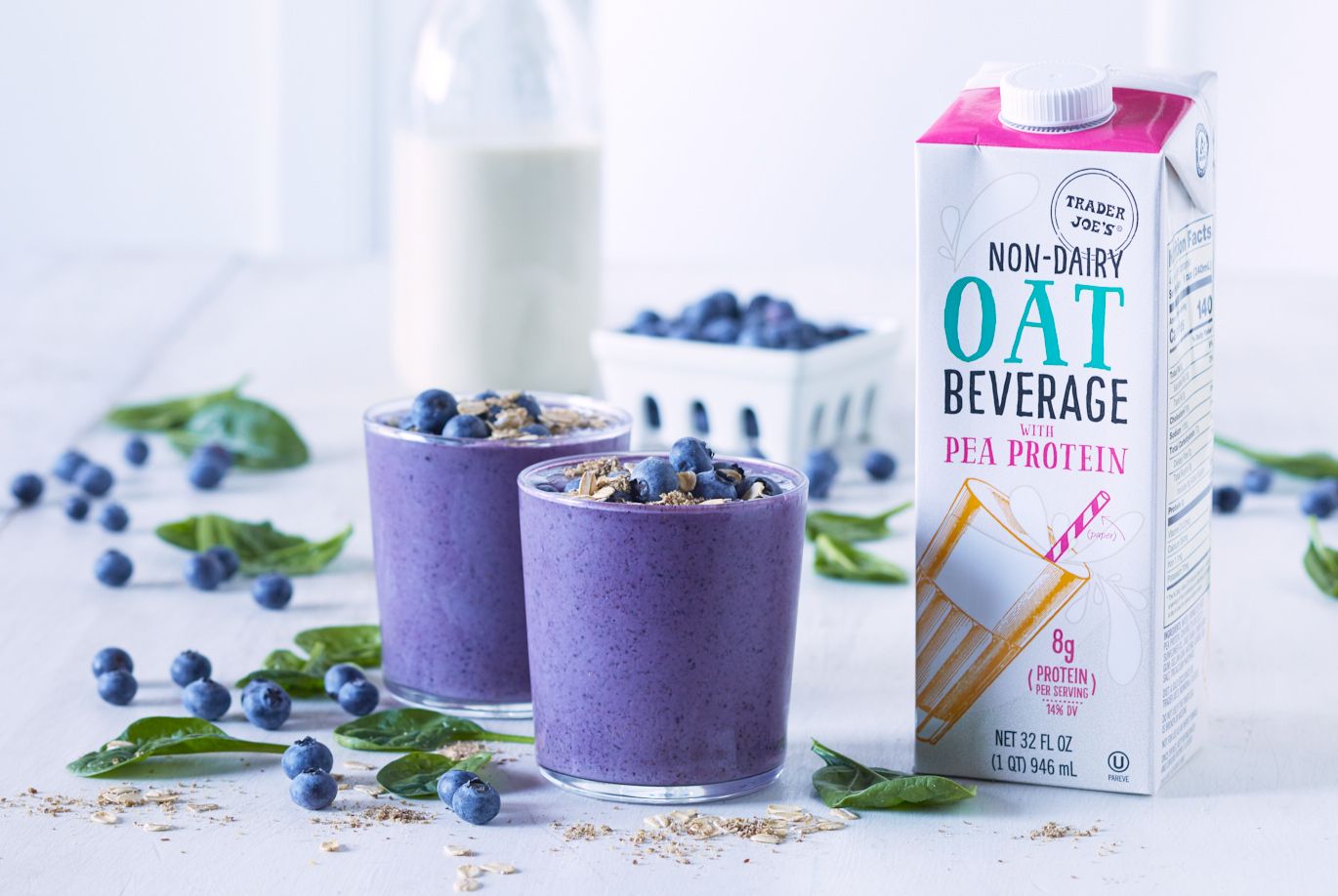 Trader Joe's Non-Dairy Oat Beverage with Pea Protein used in recipe for Blueberry Oat Protein Smoothie; in two glasses topped wtih fresh blueberries and oats; blueberries and spinach leaves on white surface